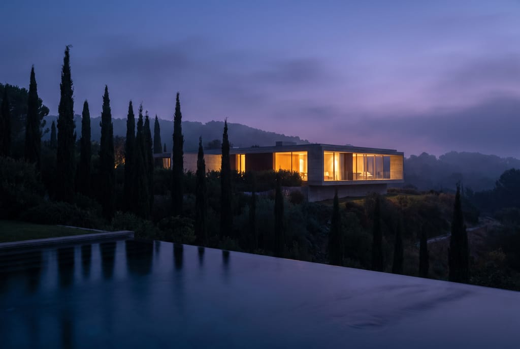 Villa contemporaine au crépuscule, intérieur baigné de lumière chaude vu à travers les baies vitrées, jardin de cyprès silhouettés en heure bleue